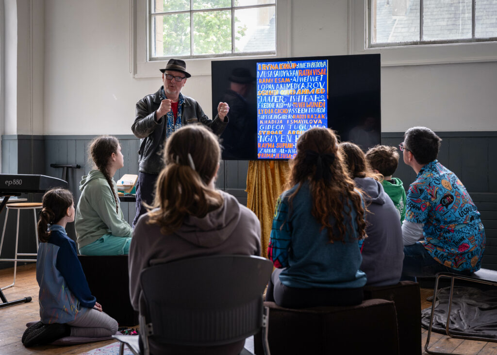 A man presenting a blue digital poster on a TV screen to a group of 7 children seated on chairs circling him.