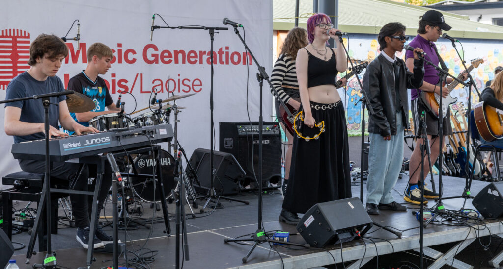 6 young people are performing on an outdoor stage in a marquee. 4 are standing playing guitar, tambourine and singing. Two are sitting playing the drums and keyboard. The sign in the background reads 'Music Generation Laois'.