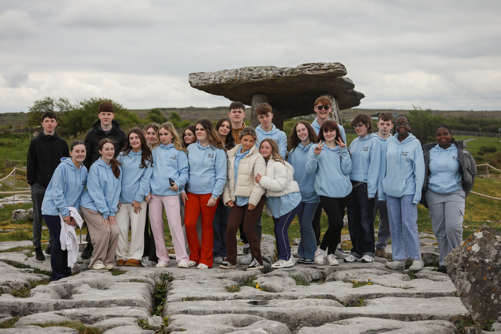 21 young people stand in front of a green landscape and a dolmen. Most of them are wearing blue 'Living Legends' hoodies.